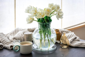 A vase of white flowers on a table beside a steaming mug of coffee.