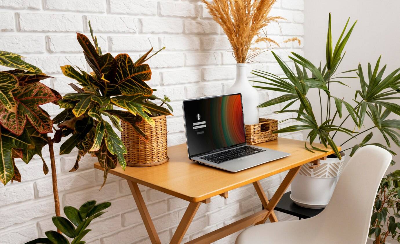A cozy home office setup featuring a laptop on a wooden desk surrounded by lush indoor plants and a white brick wall.