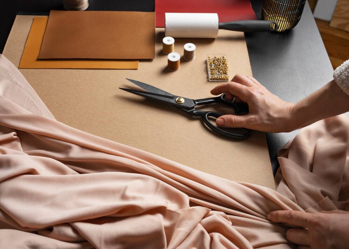 Person cutting beige fabric with scissors on a wooden table, surrounded by sewing supplies.