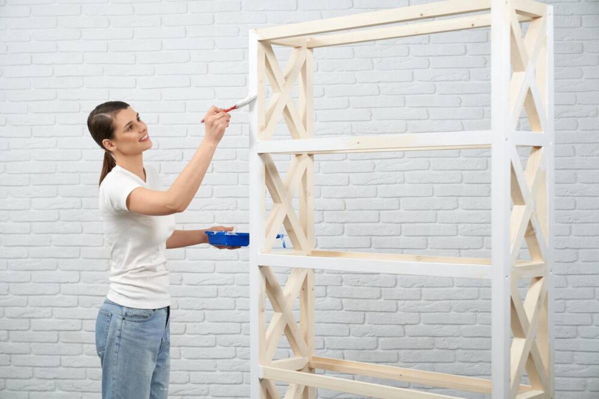 Woman painting a wooden shelf against a white brick wall.