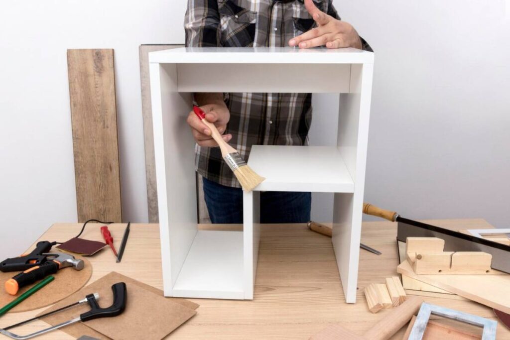 Person assembling a small white shelf with tools and woodworking supplies on a wooden table.