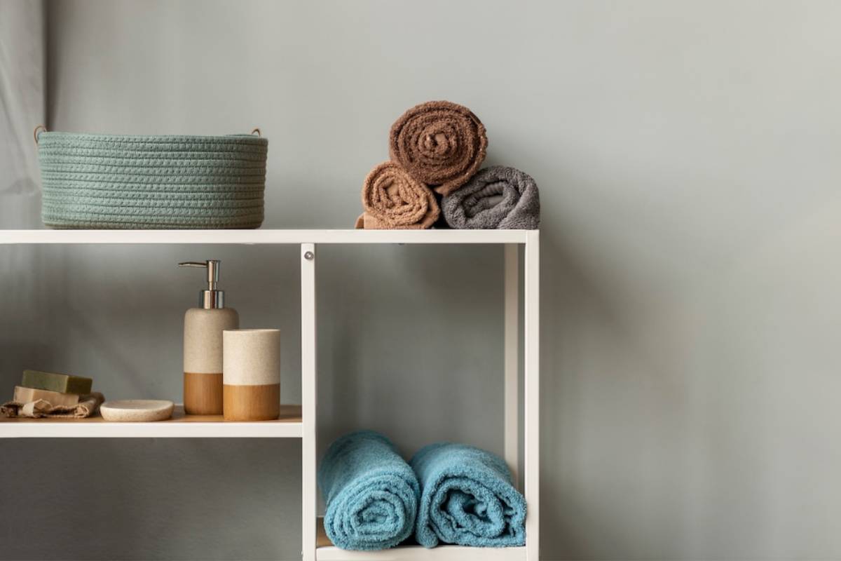 A neatly organized bathroom shelf with rolled towels, a basket, and toiletry dispensers on a muted gray background.
