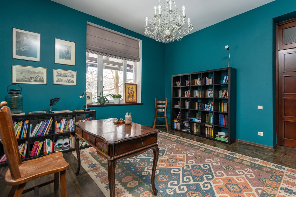 A cozy study room with teal walls, a classic wooden desk, ornate rug, bookshelves, and hanging chandelier.