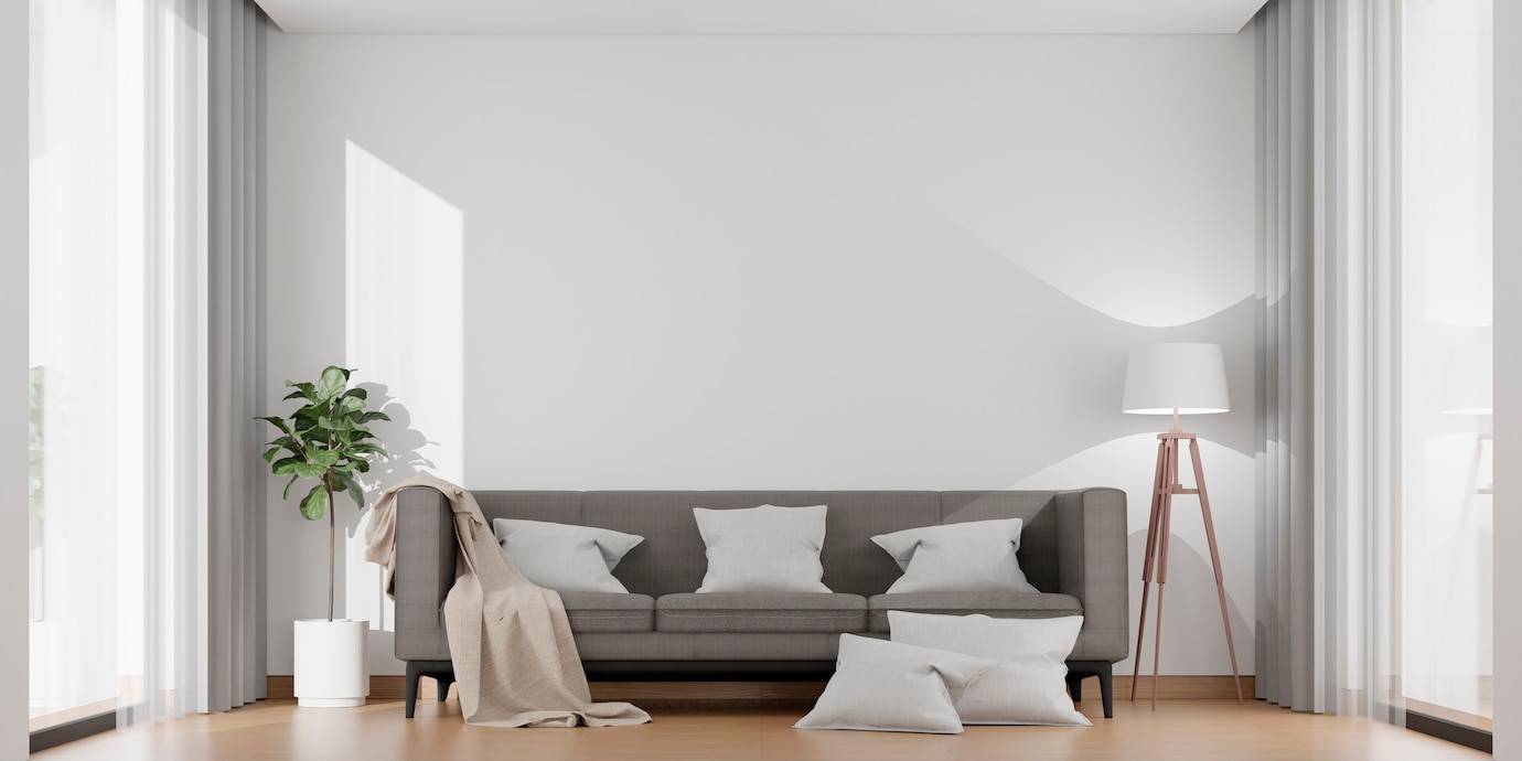 Minimalist living room with grey sofa, white pillows, floor lamp, and a potted plant by large windows.