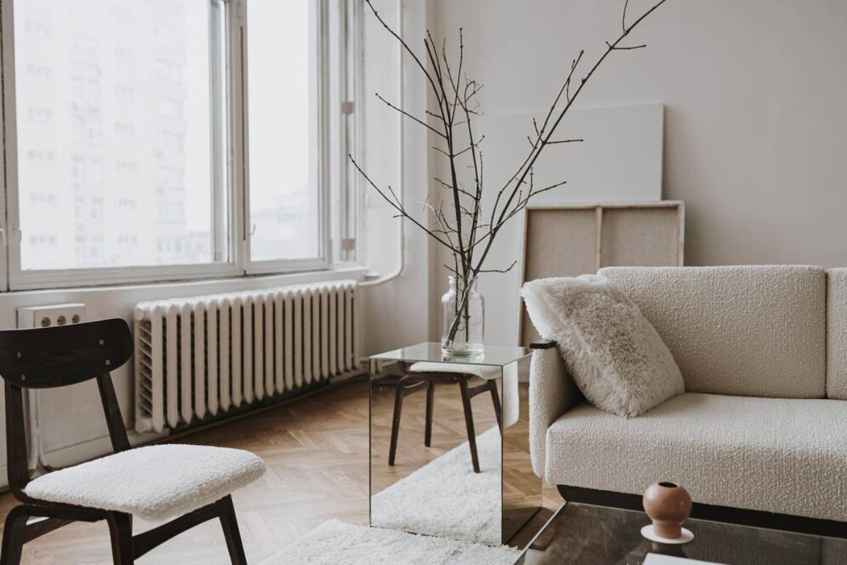 Minimalist living room with a beige sofa, glass table, and bare tree branches in a vase by large windows.