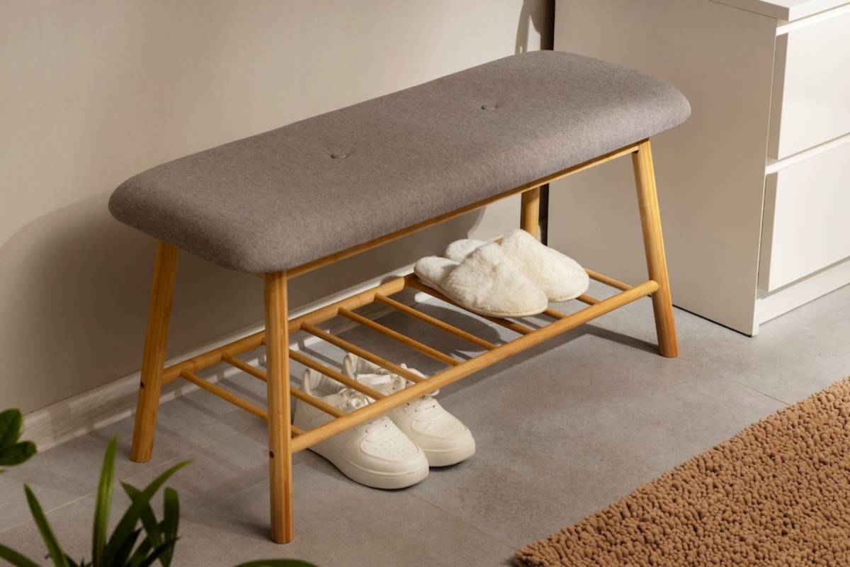 Modern gray upholstered bench with wooden legs and a shoe rack, holding white sneakers and fluffy slippers.