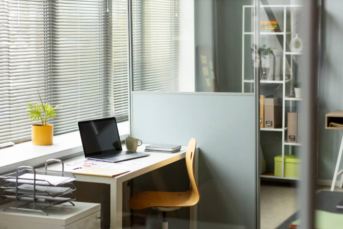 Modern office workspace with a laptop, coffee mug, and a plant on the desk, situated by blinds-covered windows.