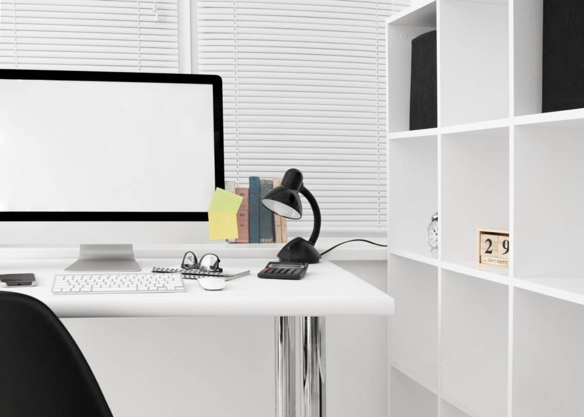 Modern home office setup with a desk featuring a computer, lamp, and neatly arranged shelves next to blinds.