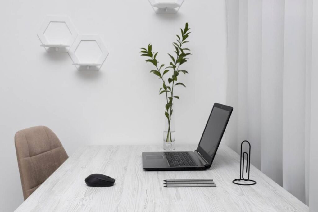 Minimalist home office setup with open laptop, mouse, and office supplies on a white table, near a potted plant and modern wall lights.