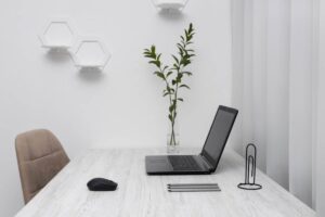 Minimalist home office setup with open laptop, mouse, and office supplies on a white table, near a potted plant and modern wall lights.