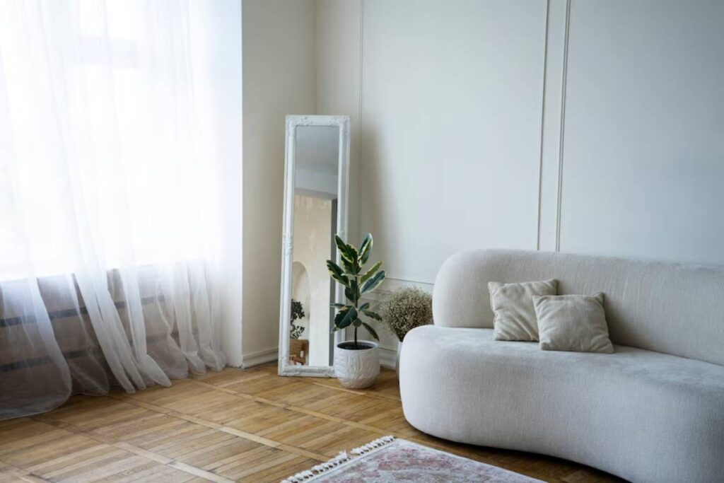 Bright living room with a white sofa, large floor mirror, and potted plant near sheer curtains.
