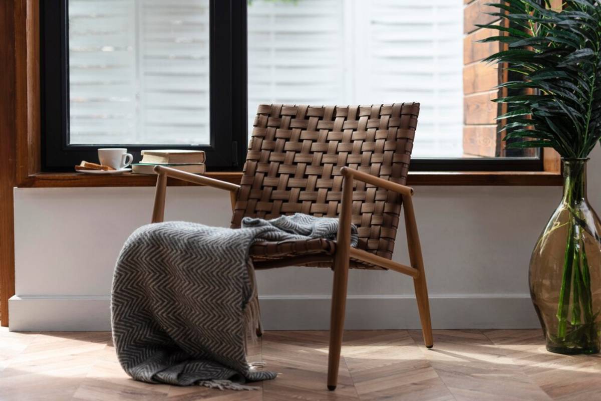 A cozy interior featuring a woven chair next to a window, with a blanket, a coffee cup, books, and a large plant.