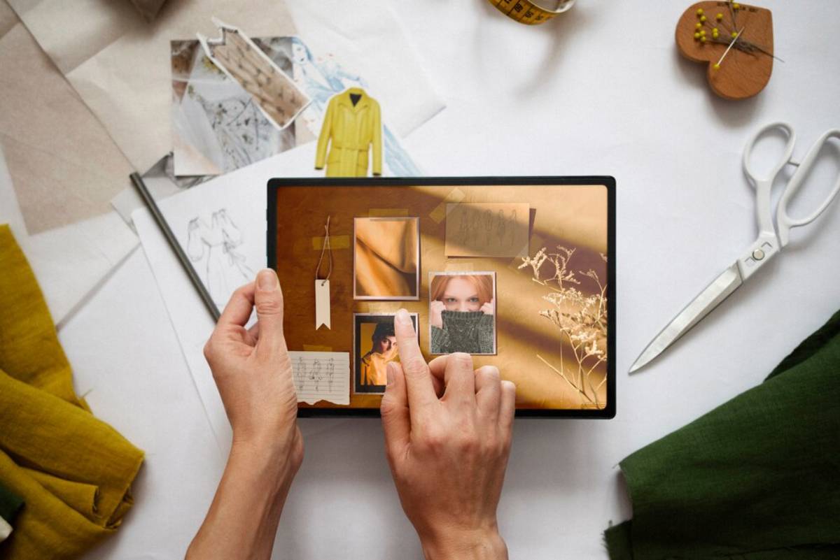 A hand interacts with a tablet displaying a collage of photos and fabric swatches, surrounded by design sketches and tailoring tools.
