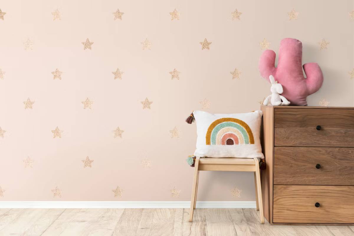 Child's room with a star-patterned wallpaper, wooden dresser, rainbow pillow on a chair, and a pink cactus plush toy.