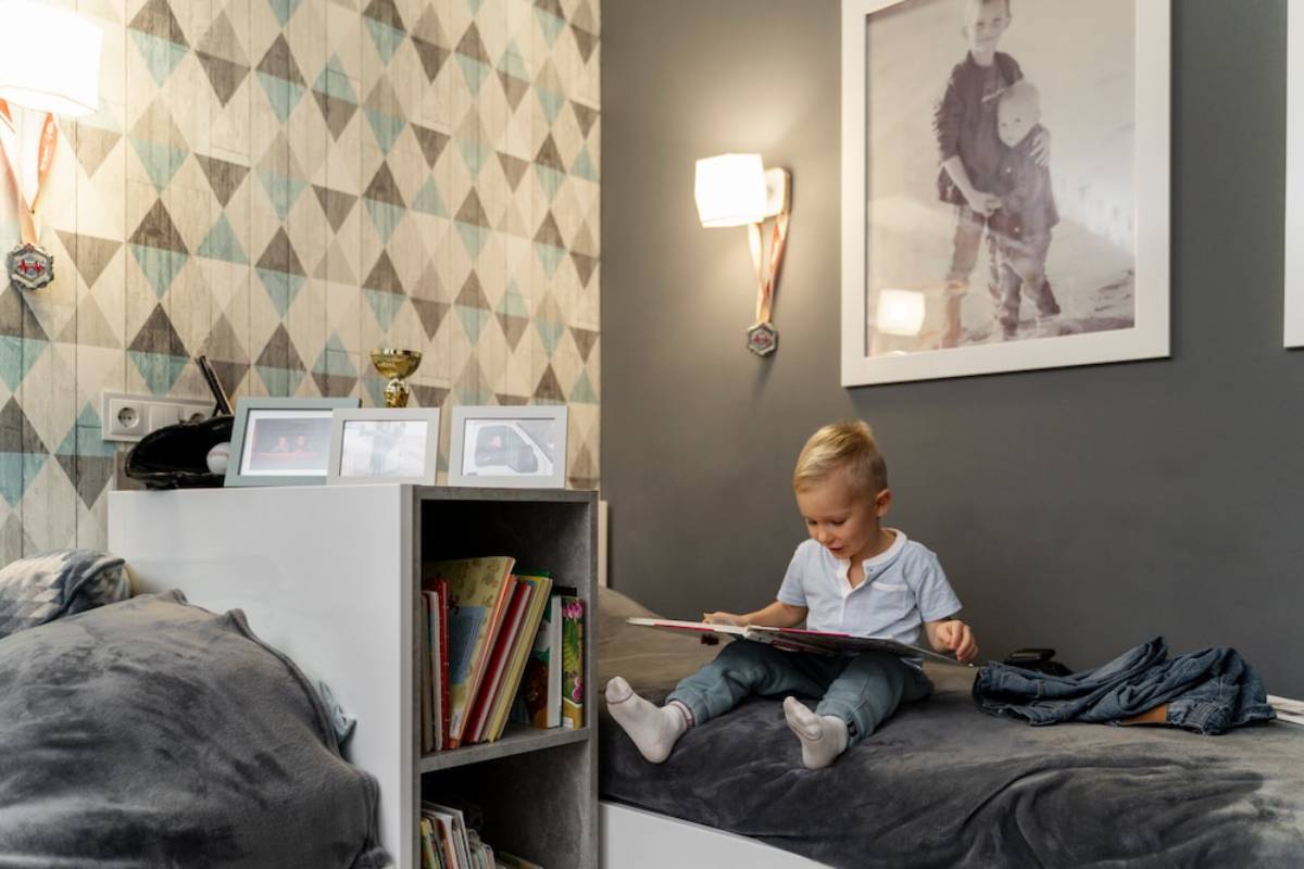 A young child reading a book in a stylish, geometrically wallpapered bedroom.