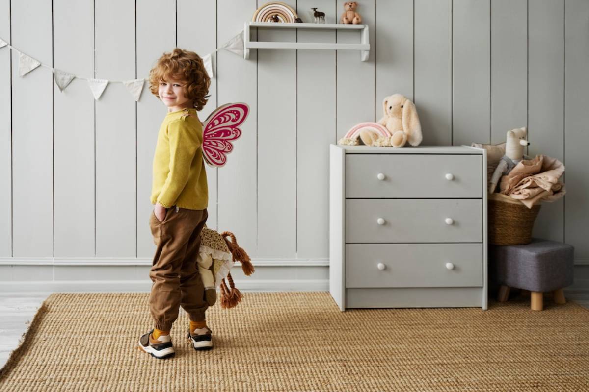 Young child with curly hair wearing pink fairy wings in a playful room, looking back over shoulder with a soft smile.