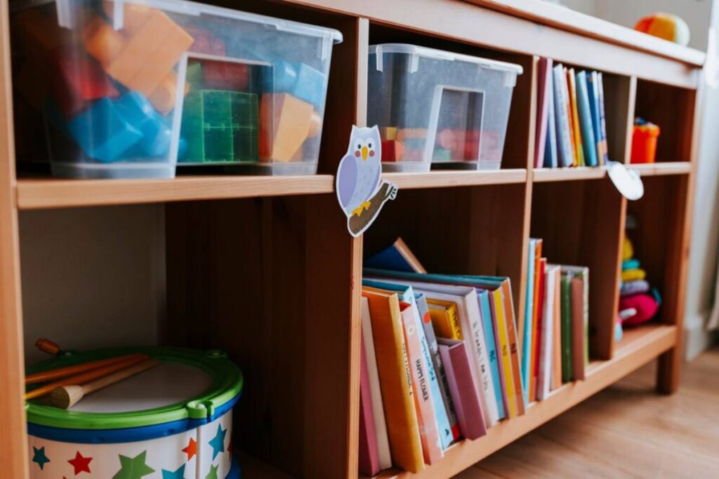 A wooden shelf storing children's books, toys in clear bins, and a colorful drum, adorned with a cute owl sticker.