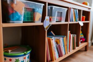 A wooden shelf storing children's books, toys in clear bins, and a colorful drum, adorned with a cute owl sticker.