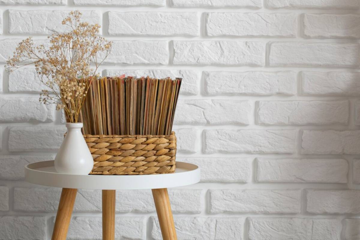 A small table with dried flowers in a vase and a wicker basket full of magazines against a white brick wall.