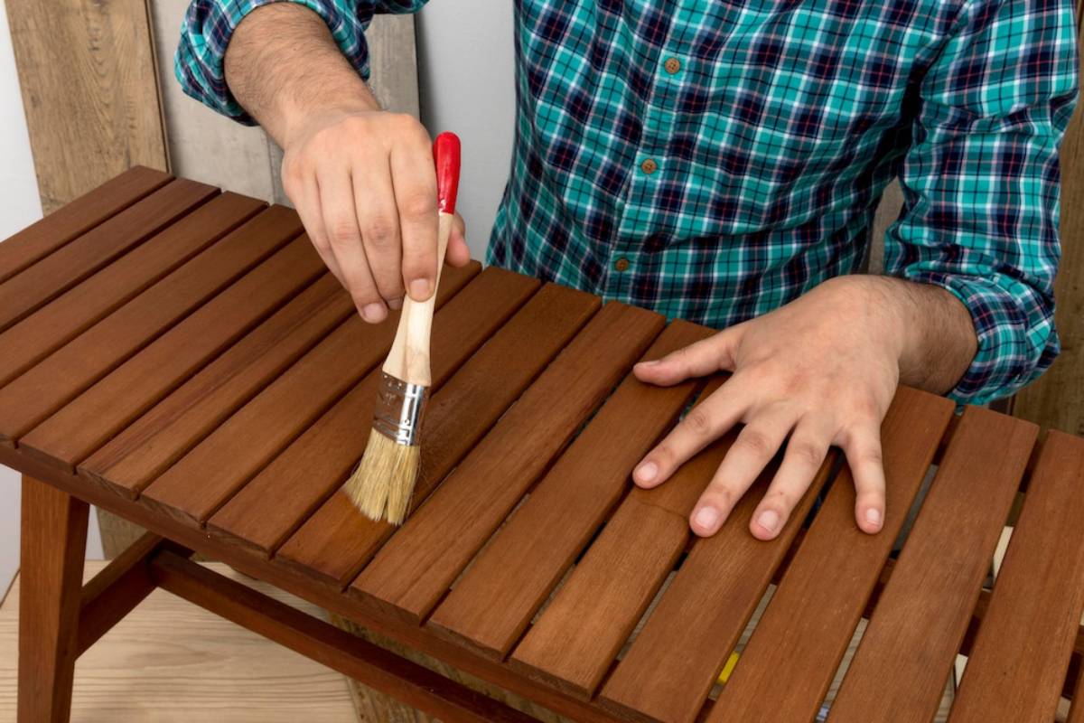 A person in a plaid shirt is using a brush to apply finish on a wooden slatted table.