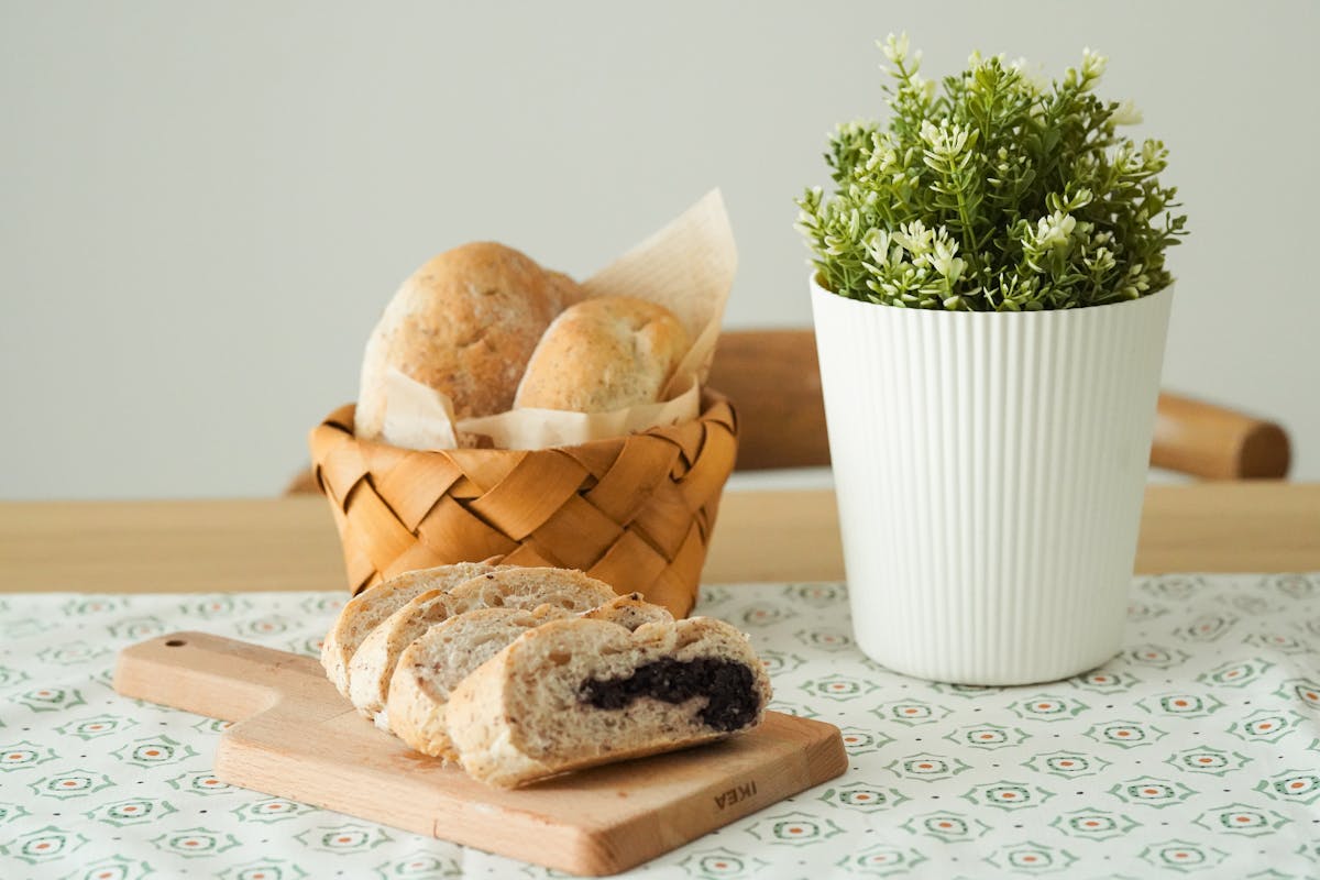 A wooden cutting board displays sliced bread with a dark filling, next to a basket of rolls and a potted plant